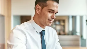 Person in a white shirt and tie smiling while working at a desk.