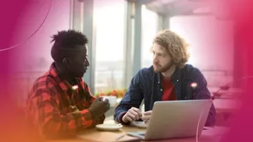Two people having a discussion at a table with a laptop and notepads.