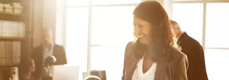 A person smiling in a sunlit office with colleagues working in the background.