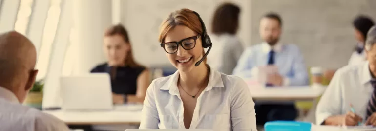 Person with headset smiling at computer in a busy office.