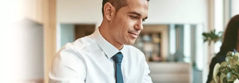 Person in a white shirt and tie smiling while working at a desk.