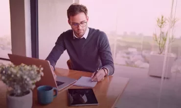 Person working at a desk with a laptop and notepad near a window.